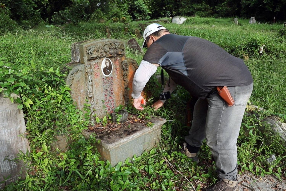 Mr Peter Pak snipping away with shears at overgrown grass and roots over a tombstone in Bukit Brown Cemetery on June 18, 2022. — TODAY pic