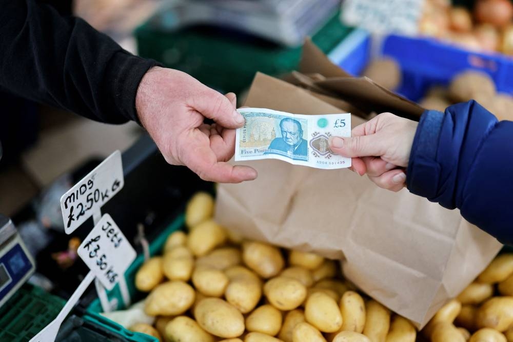In this file photo taken on November 21, 2021 A customer (R) pays passes a stallholder a five pound note to pay for their fruit and vegetables at Walthamstow Market in east London. — AFP pic