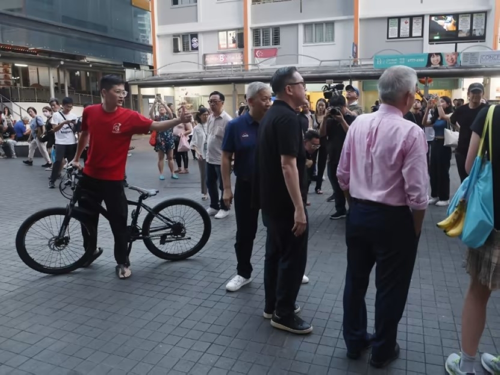 A cyclist (far left) creating a scene when presidential candidate Ng Kok Song (right, back facing camera) was greeting members of the public outside Clementi Mall during a walkabout on Aug 29, 2023. — TODAY pic