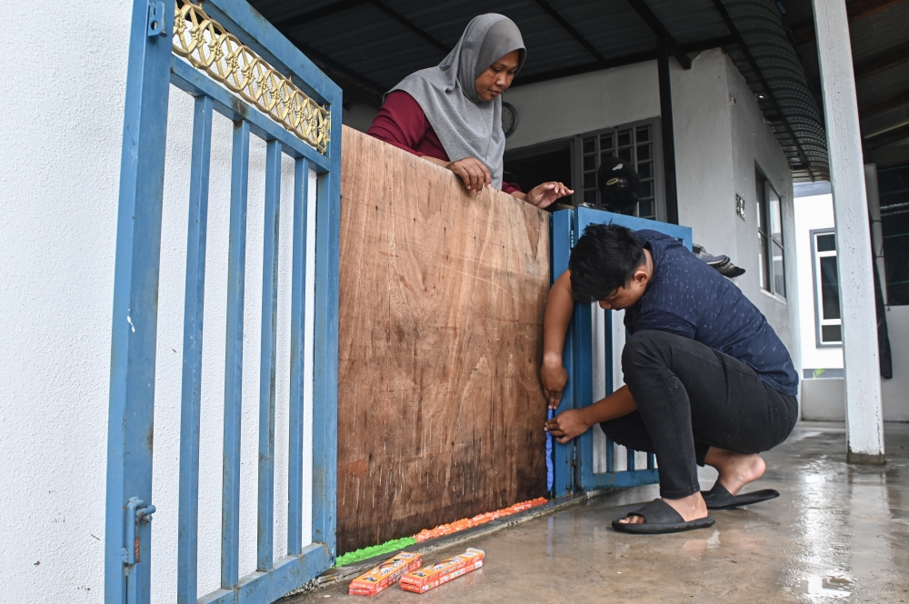 Norlida Ramli (left) watches as her son Wan Azrol Haikal Wan Azmadi sticks plasticine to gaps around the plywood walls installed on the front and back fences of their house in Kampung Padang Gong Pak Maseh, Terengganu November 23, 2023. — Bernama pic