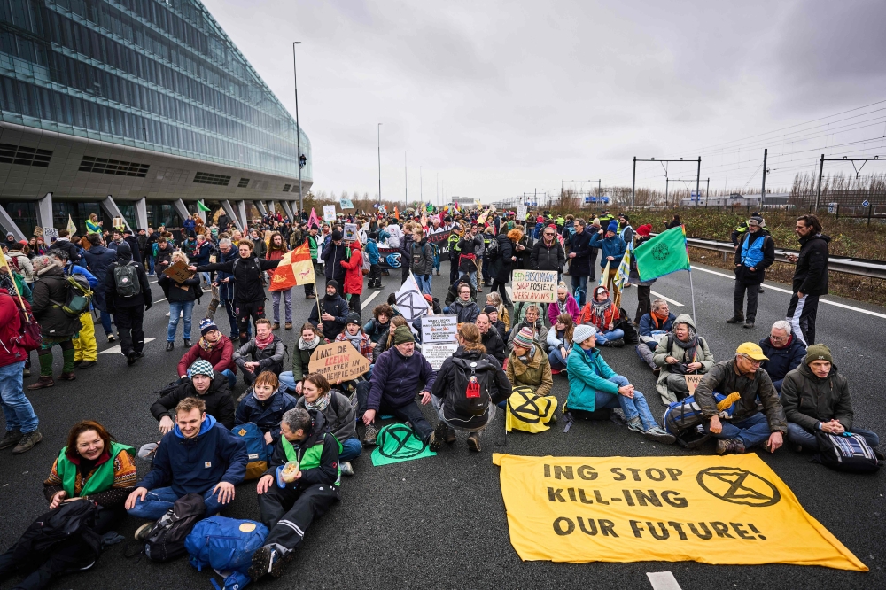 Protesters sit by a banner reading ‘ING Stop Kill-ING Our Future!’ during a blockade action of the A10 highway by enviromentalist group Extinction Rebellion in front of the former headquarters of Dutch multinational banking and finance corporation, ING to protest against the bank's financing services to the fossil industry, in Amsterdam on December 30, 2023. — AFP pic