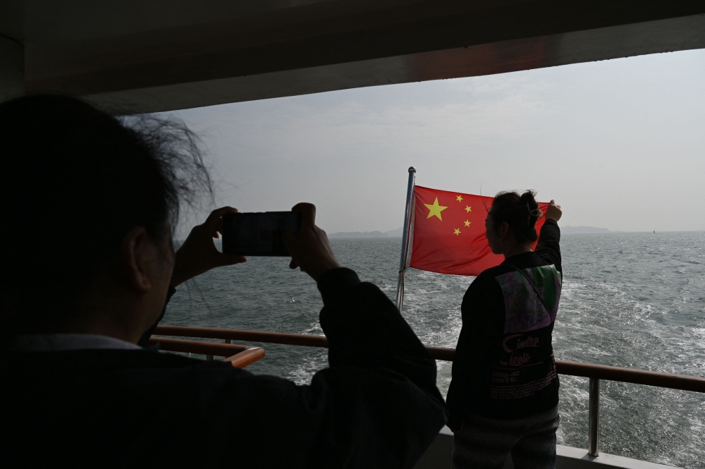 A Chinese man sings as he holds the the Chinese flag, with the backdrop of Taiwanese islands from a distance of 2 kilometres, during a boat tour to view the islands, off Xiamen, in China’s south-east Fujian province on January 11, 2024, two days before Taiwan’s Presidential election. — AFP pic
