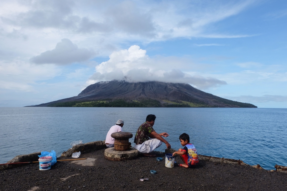 People fish at the port on Tagulandang island in Sitaro, North Sulawesi on April 19, 2024, as the Mount Ruang volcano is seen in the background spewing smoke. — AFP pic