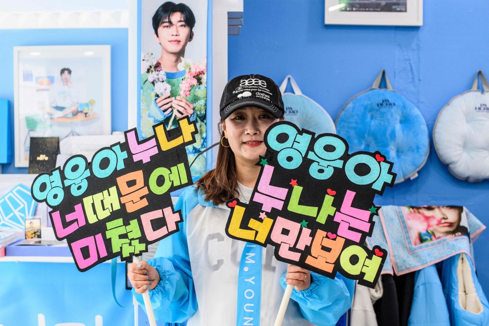 This picture taken on June 3, 2024 shows Shin Ji-min, a fan of South Korean trot singer Lim Young-woong, posing under a photo of Lim while holding signs reading 'Young-woong you make Noona go crazy, Young-woong Noona can only see you' at a sky-blue cafe named ‘Woong's Tree’, which stands as a shrine to the singer, in his hometown of Pocheon city. — AFP pic