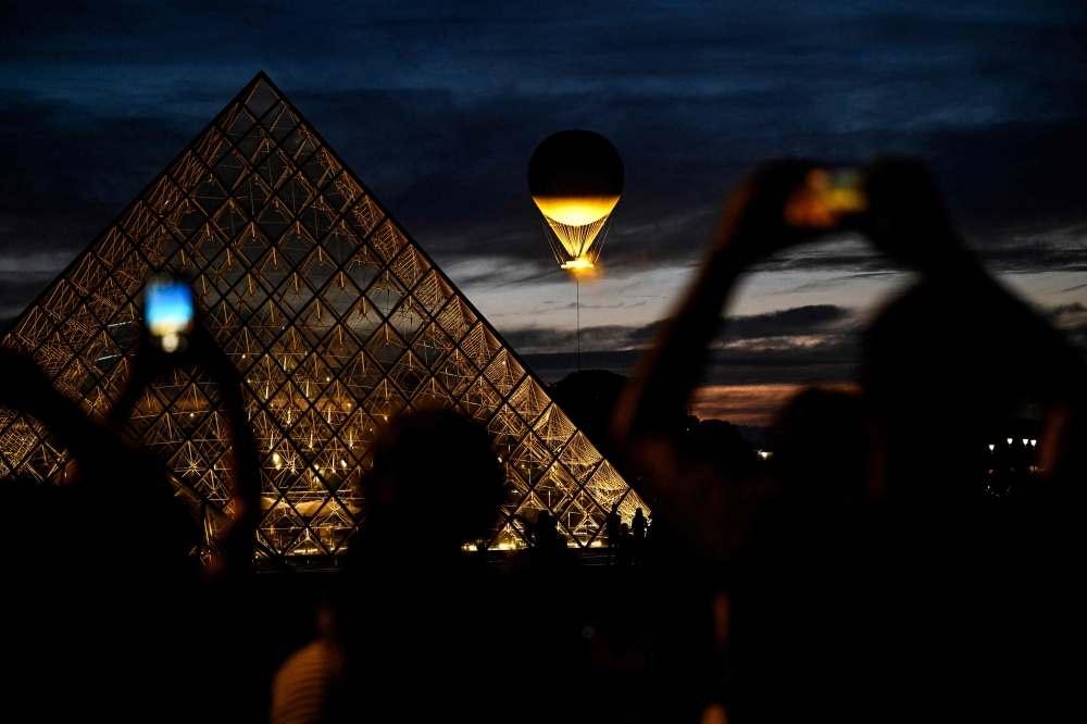 Bystanders take photographs of the Paris 2024 Olympic Games cauldron attached to a balloon overlooking the Louvres glass pyramid designed by Chinese-US architect Ieoh Ming Pei in Paris on August 6, 2024. — AFP pic