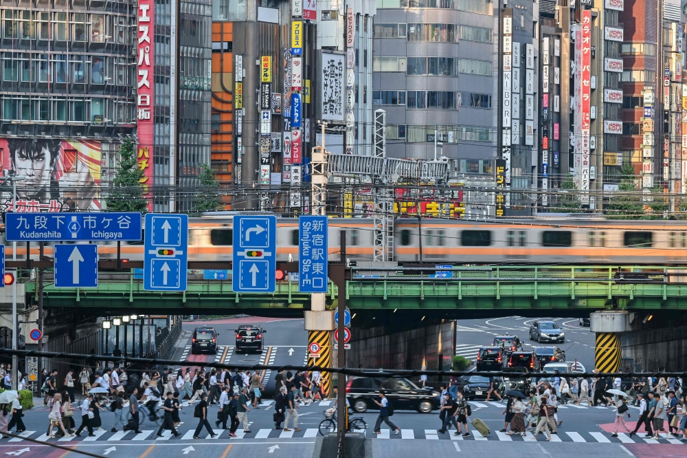 Pedestrians cross an intersection as a train goes by overhead, in the Shinjuku area of central Tokyo on August 13, 2024. — AFP pic