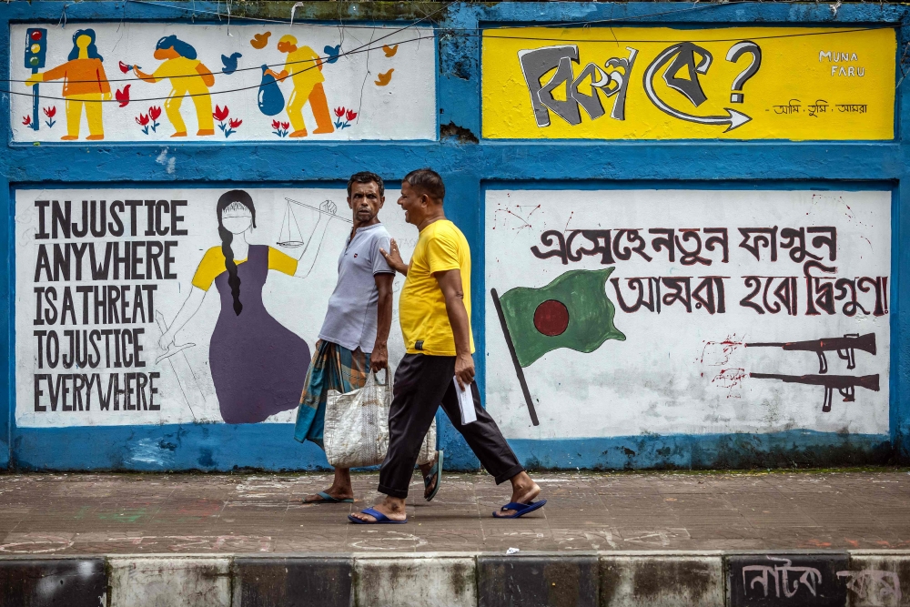 Two men walk in front of a wall decorated in support of the students movement after the resignation of Sheikh Hasina, August 18, 2024. — AFP pic