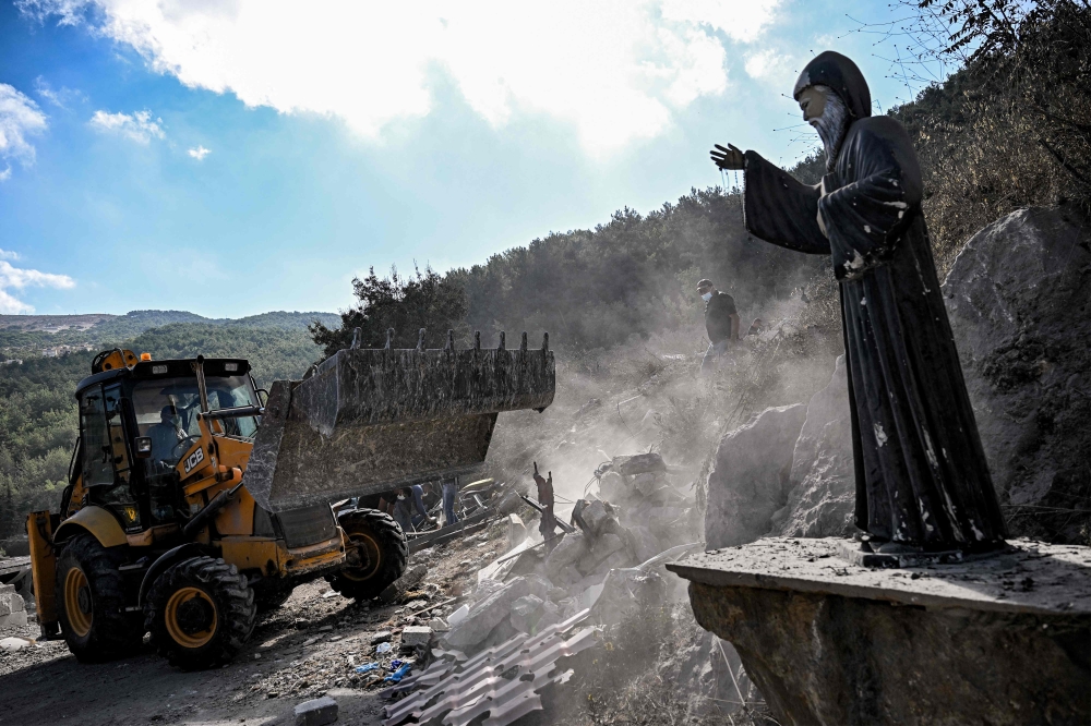 A Statue of the 19th-century Maronite Christian saint Mar Charbel is pictured as a bulldozer moves to clear rubble and debris from the site of a previous Israeli air strike on the village of Aito in northern Lebanon on October 15, 2024. — AFP pic
