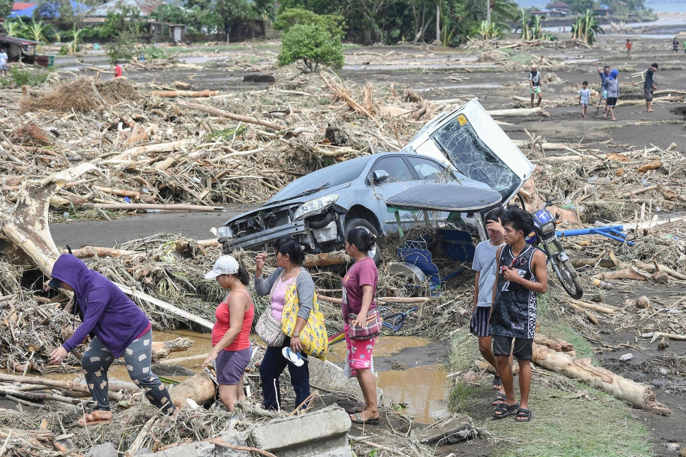 People walk past destroyed vehicles swept away along with debris of logs due to heavy rains brought about by Tropical Storm Trami in Laurel, Batangas province, south of Manila on October 25, 2024. 