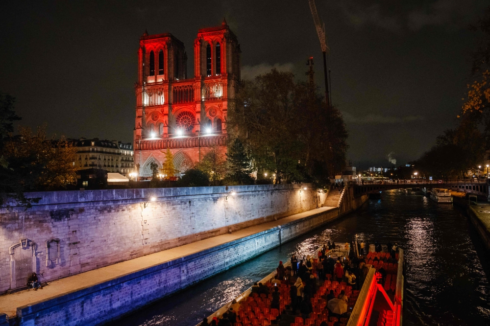 This photograph shows an open excursion boat (Bateau-Mouche) passing in front of the Notre-Dame cathedral illuminated in red, as part of the 