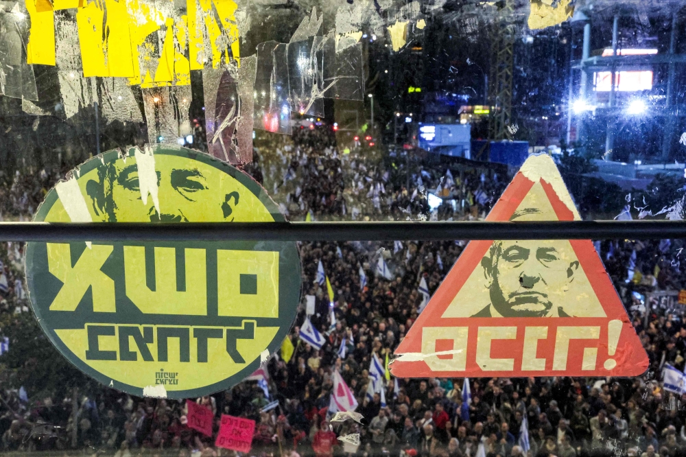 Stickers critical of Israeli Prime Minister Benjamin Netanyahu are pictured behind the glass of a window above demonstrators gathering outside Israeli Defence Ministry headquarters Tel Aviv on March 22, 2025. — AFP pic
