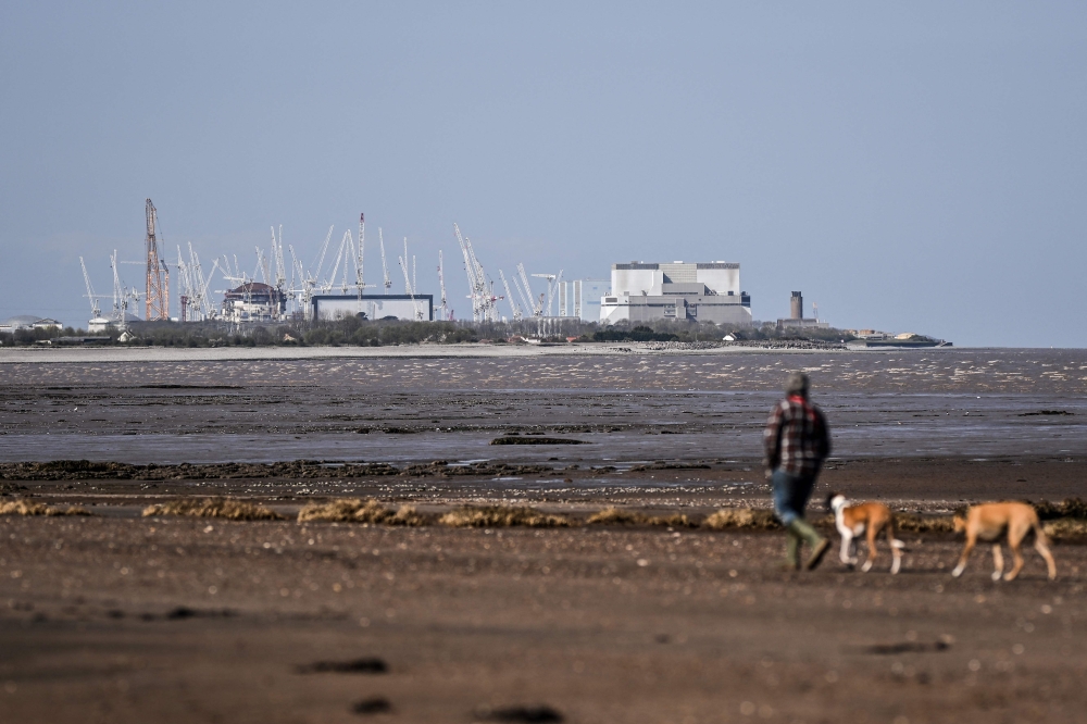 A person walks their dogs by the water at the WWT Steart Marshes, in Bridgwater, Somerset, western England, on April 5, 2025, with the Hinckley point nuclear power station in the background. Britain has moved the Hartlepool nuclear power station in north-east England operated by France’s EDF into enhanced regulatory attention for safety. — AFP pic