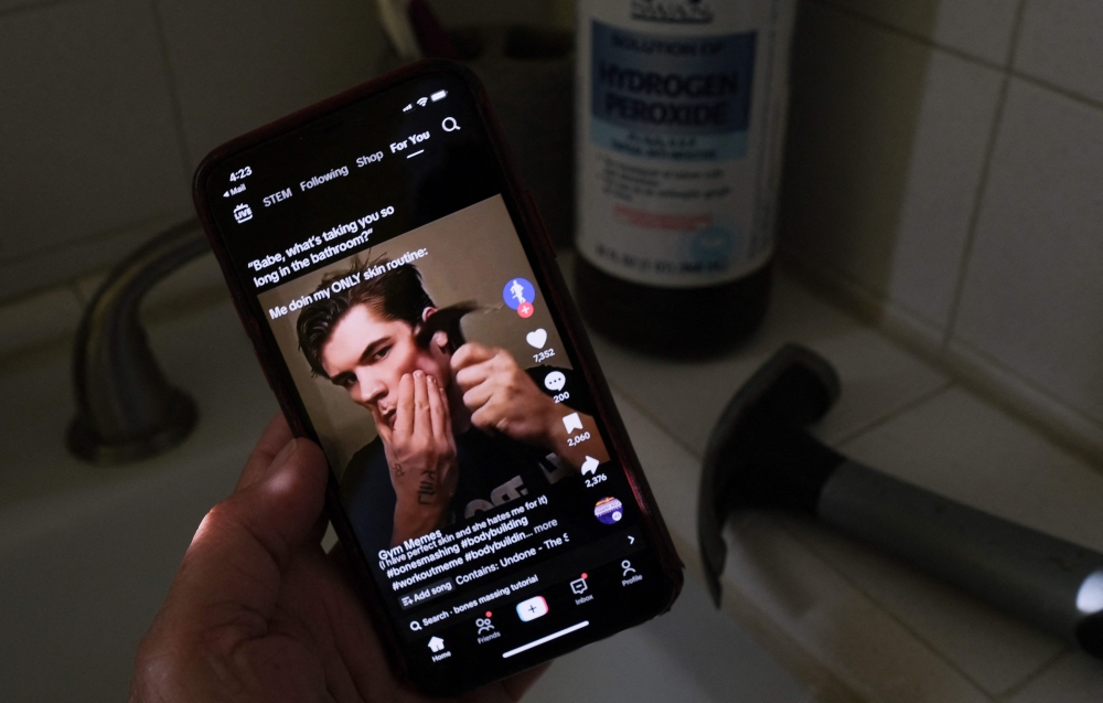 This illustration photo shows a video of male TikTok influencer hammering his cheekbone, playing on a smartphone in a bathroom next to a bottle of hydrogen peroxide and a hammer in Los Angeles. — AFP pic