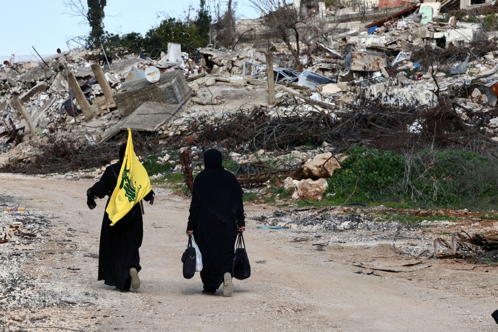 Women walk near destroyed buildings, with one holding the flag of Hezbollah, in the southern Lebanese village of Kfar Kila. The group's new leader Naim Qassem has vowed not to disarm despite mounting Israeli and US pressure. — Reuters pic