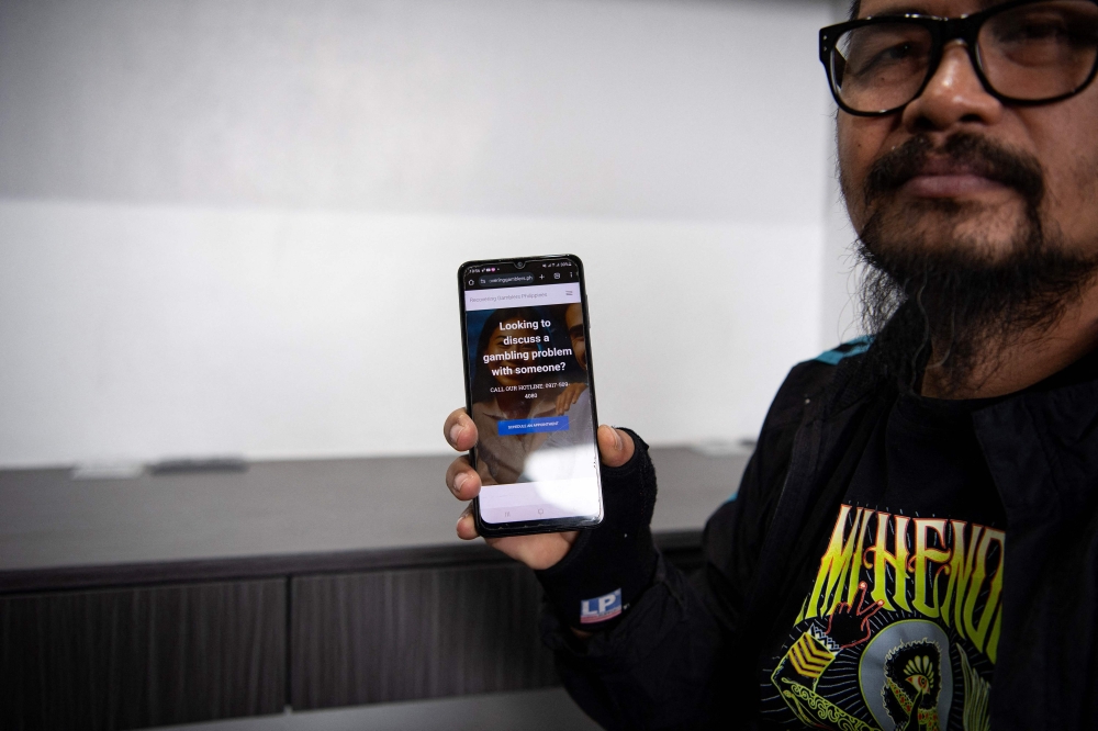 Ray Gibraltar, former film and TV director-turned-painter holds a mobile phone showing the website of the Recovering Gamblers of the Philippines, founded by Reagan Praferosa, in Manila July 22, 2025. — AFP pic