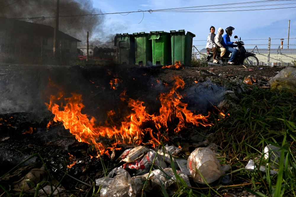 This photo taken on January 30, 2025 shows people travelling on a motorbike past burning plastic waste on the outskirts of Phnom Penh. — AFP pic