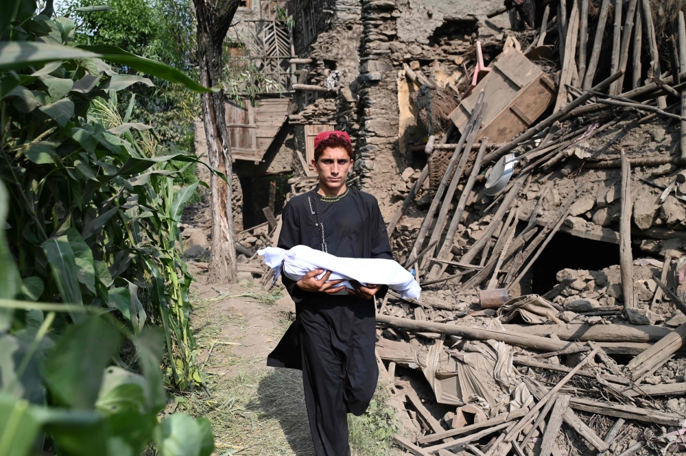 An Afghan boy carries the body of a relative in a shroud as he walks past by a damaged house following earthquakes in the Mazar Dara village of Nurgal, a district of the Kunar Province, in Eastern Afghanistan, on September 1, 2025. — AFP pic