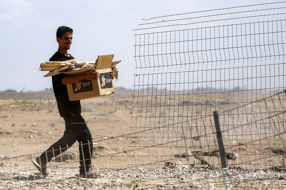 A man walks with a cardboard box bearing the Gaza Humanitarian Foundation (GHF) logo and loaded with pieces of wood as he walks near a fence that was a barrier at the so-called 