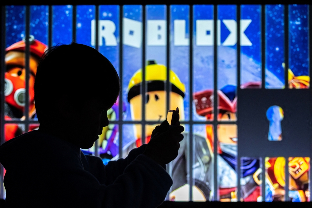A boy poses for a photo while holding a smartphone in front of a screen displaying characters of the US children's video game Roblox, in this illustration taken December 8, 2025. Roblox, which reported some 112 million daily users worldwide in the second quarter of this year, recently tightened its controls to protect minors. — AFP pic 
