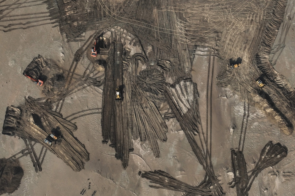 An aerial photograph shows excavators and bulldozers clearing sand dunes and extracting clay in the deser