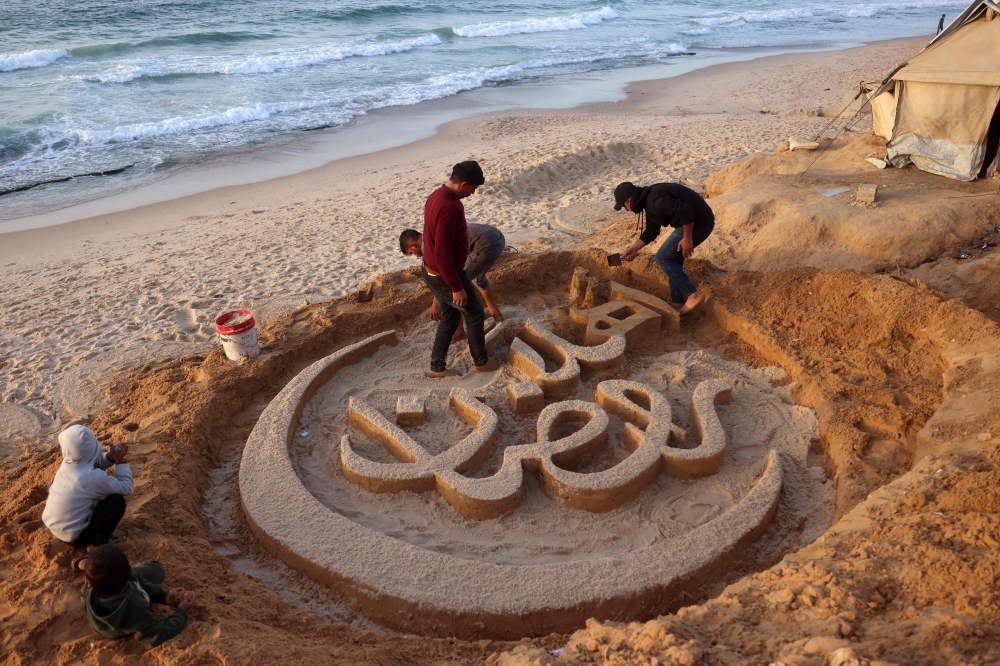 People assist Palestinian artist Yazeed Abu Jarad, who was displaced by the war from Beit Lahia in the northern Gaza Strip, as he creates a sand sculpture with the greeting message 