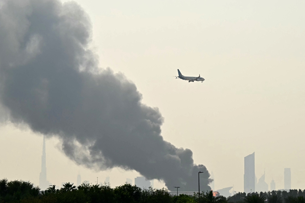 An Emirates aircraft flies past plumes of smoke from an ongoing fire near Dubai International Airport in Dubai on March 16, 2026. — AFP pic