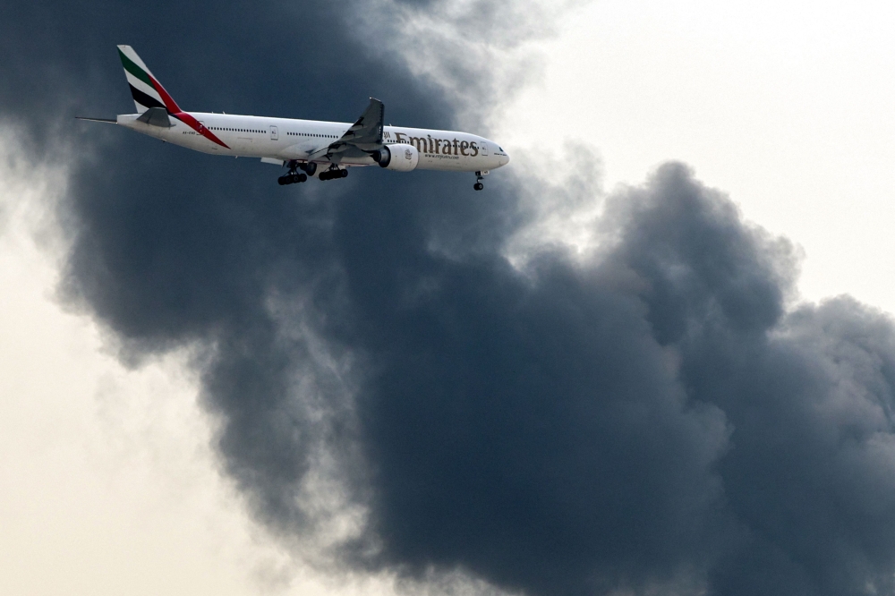 An Emirates Boeing 777 aircraft prepares for landing as a smoke plume rises from an ongoing fire near Dubai International Airport in Dubai on March 16, 2026. — AFP pic