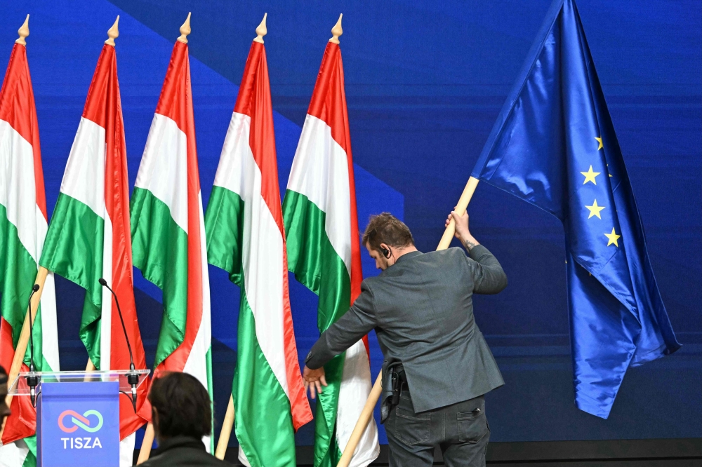 A staff member adjusts an EU flag beside Hungarian flags at the HUNGEXPO Congress and Exhibition Center in Budapest on April 13, 2026. — AFP pic