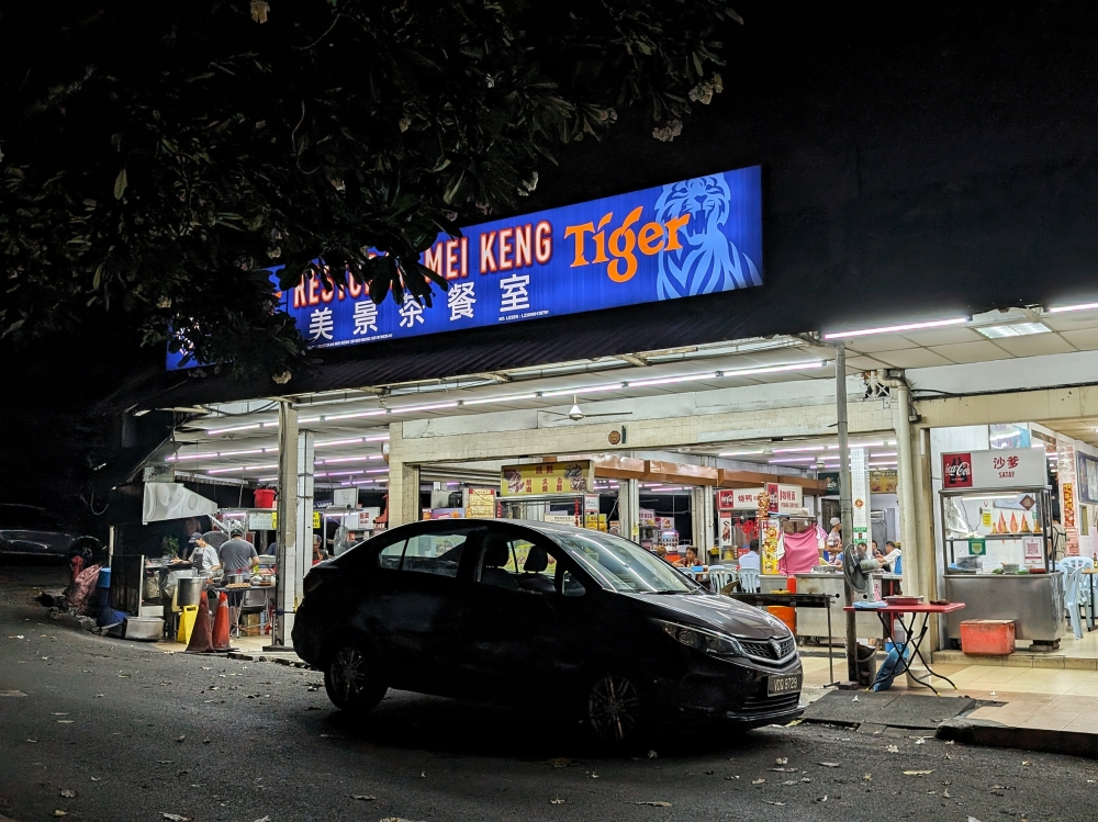 The tree slightly obscures Restoran Mei Keng’s sign, but the coffeeshop’s glaring white lights are impossible to miss at night. — Picture by Ethan Lau