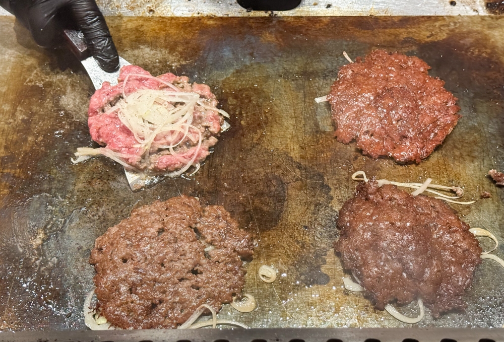 The smashed patties topped with sliced onions and cheese are cooked on the hot griddle. 