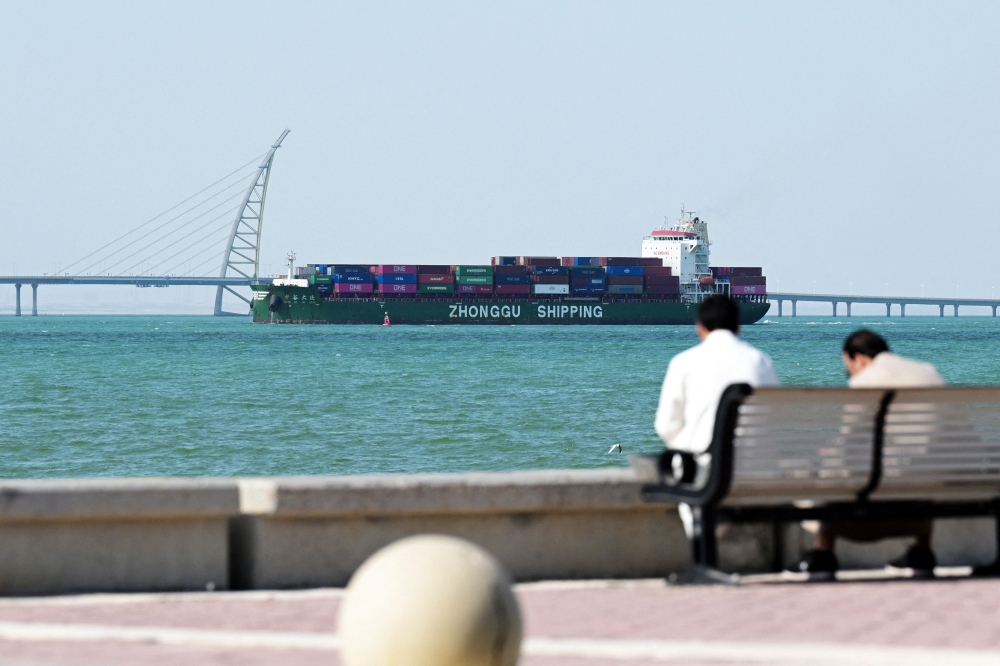 People sit on a bench on the Sheikh Jaber al-Ahmad al-Sabah Causeway as a Zhonggu Shipping container ship sails past on its way towards the port of Shuwaikh in Kuwait City on February 28, 2026. — Reuters pic 