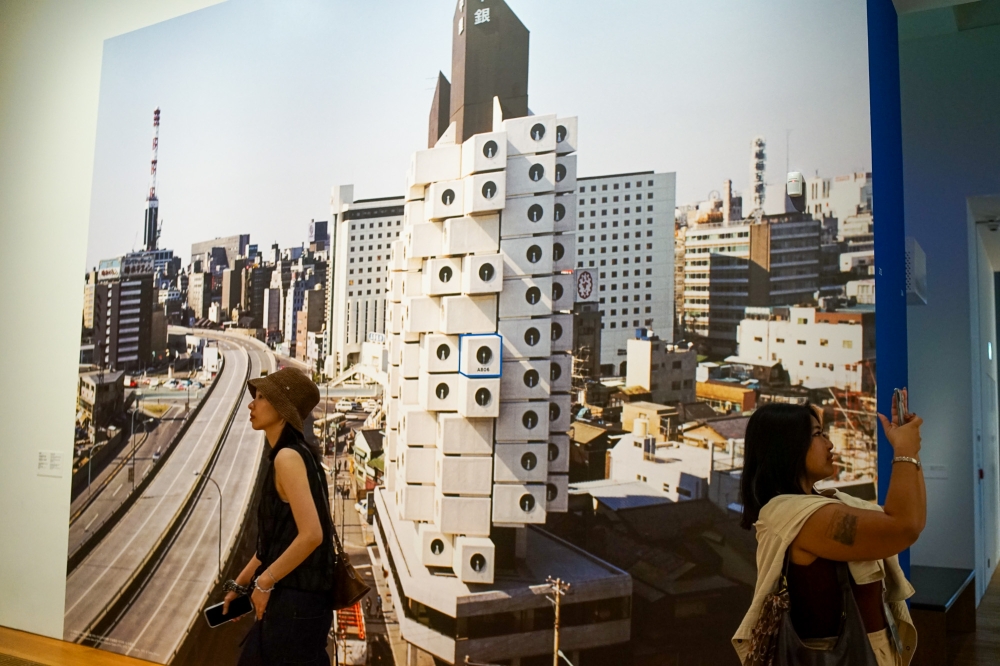 Visitors at M  walk past a photograph of the Nakagin Capsule Tower, with a box marked with blue outlines being Unit A806’s location in the building which has since been demolished. — Picture by Ida Lim