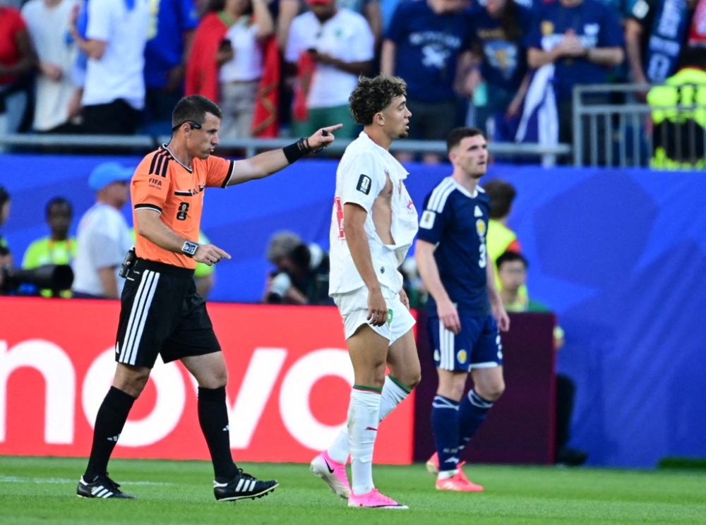 Morocco midfielder Neil El Aynaoui shows referee Ilgiz Tantashev his torn Puma jersey during the World Cup group-stage match against Scotland, one of four high-profile shirt failures involving the manufacturer’s kits at the tournament. — AFP pic