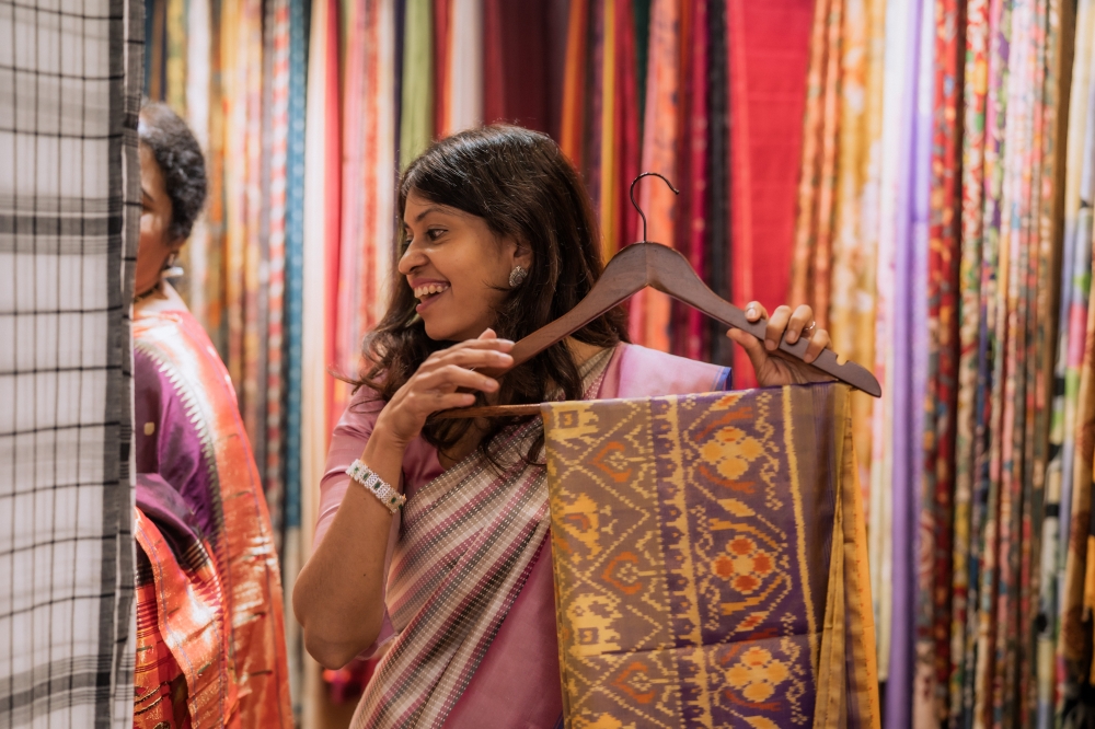 Suvitha holds up a vibrant handloom saree in Pattushastra’s Artisan Room, where every piece is made by single mothers and women from low-income communities in Kolkata. With no two designs alike, each saree carries its own story, and proceeds go to an NGO shelter that passes the earnings on to the women who made them.