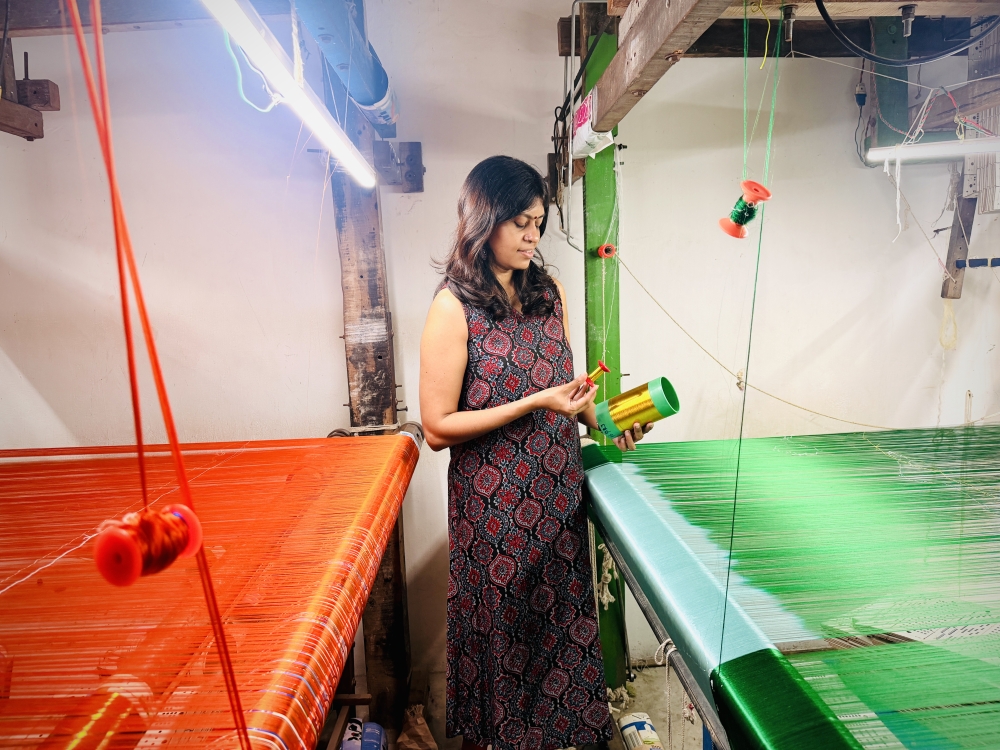 Suvitha stands between two traditional handlooms during a visit to Kanchipuram, the temple town in Tamil Nadu that gave Kanjivaram silk its name. She travels to meet the master weavers in person and checks every stage of the weaving process to make sure each saree meets the brand’s standards before it reaches Malaysia.