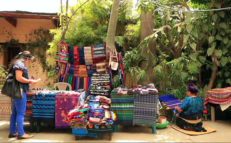 A screen capture of the video shows a tourist looking at handmade items on display at Nim Pot Market in Antigua, Guatamala.