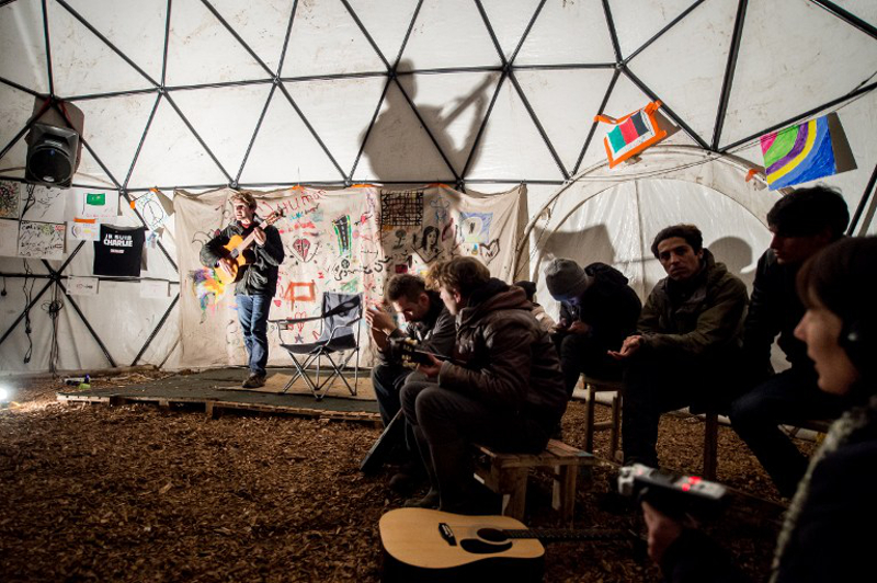 Migrants listen to a guitarist performing in a makeshift theatre, on November 25, 2015, in the so-called ‘Jungle’ migrant camp in Calais, northern France. — AFP pic