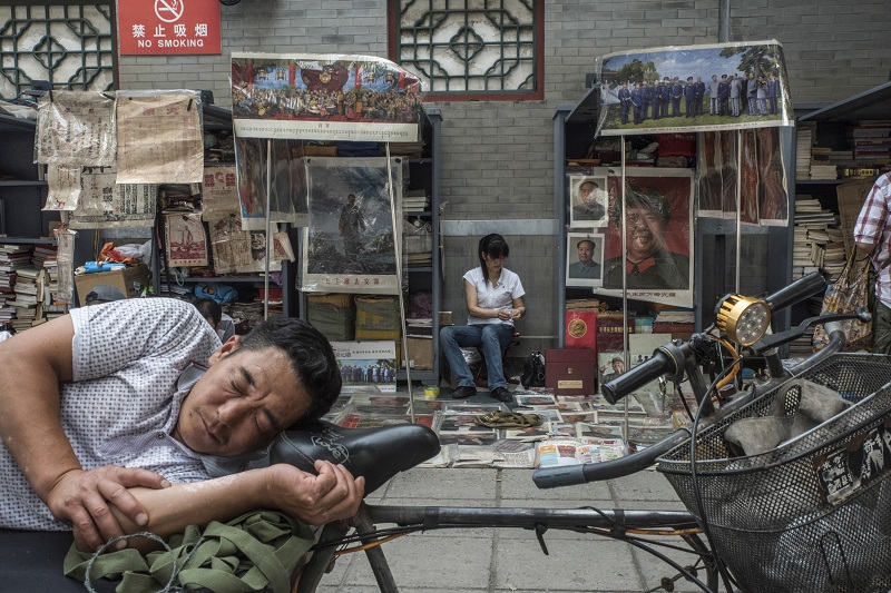 A delivery worker naps on his cart next to a stall selling Mao-era memorabilia at the Panjiayuan Antique Market in Beijing, July 9, 2016.u00e2u20acu201d Picture by Gilles Sabrie/The New York Times
