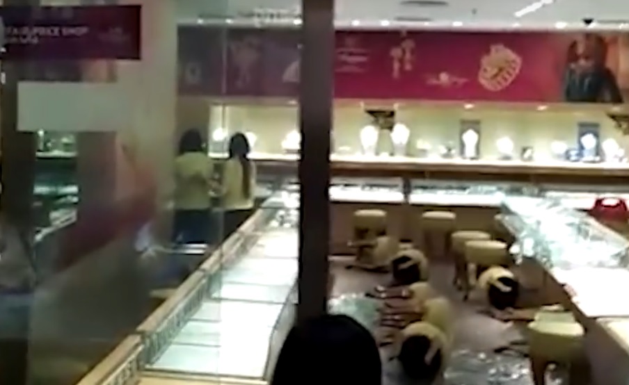 An employee comforting her colleague with a pat on the back, surrounded by smashed glass displays and upturned seat couches.