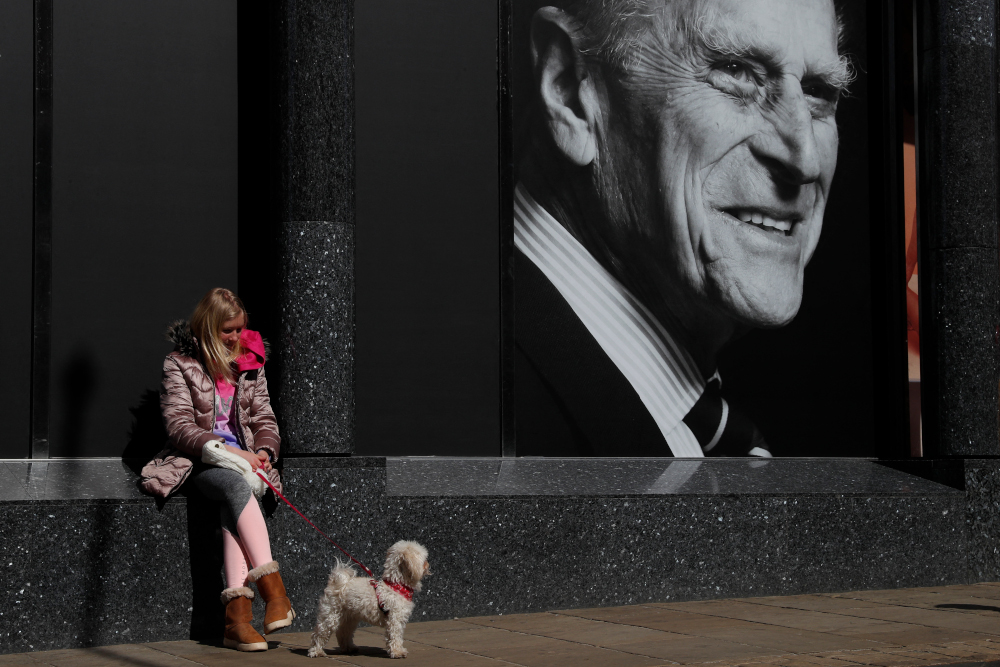 A woman and her dog sit outside a supermarket with a poster featuring Britainu00e2u20acu2122s Prince Philip, Duke of Edinburgh in the window, in the High Street of Windsor, west of London, April 16, 2021. u00e2u20acu201d AFP picnn