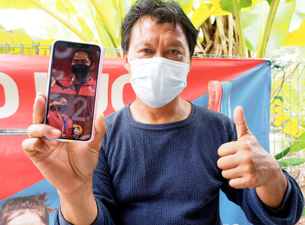 Ahmad Janius Abdullah holds a phone with photos of Olympics medallist Hidilyn Diaz at his home in Kampung Kesang Tua, Melaka, July 27, 2021. u00e2u20acu201d Bernama pic 