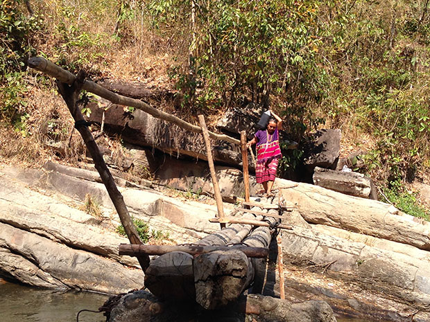 A Karen woman, dressed in a traditional outfit of handwoven fabric, makes her way carefully across a bamboo bridge.