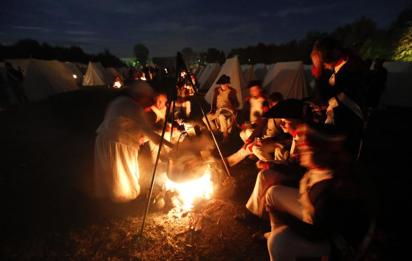 Performers wearing 19th century French military uniform have dinner next to a fire at a bivouac camp.