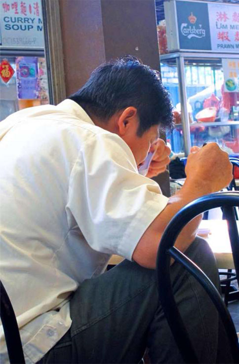 A Brickfields dining etiquette instructor demonstrating the “Two Fists with Raised Knee” approach to eating a bowl of noodles.